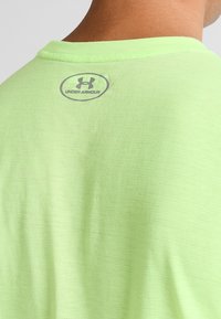 Camisa de manga corta de color verde claro con cuello redondo. Presenta una textura sutil y un logo gris de "Under Armour" en la parte trasera cerca del cuello.