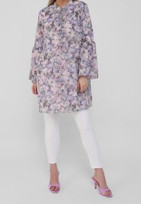 Tunique florale dans des nuances de violet et de rose, manches longues avec poignets volantés, boutonnée à l'avant, portée avec des leggings blancs et des talons violets.