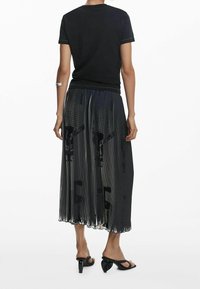 Černé tričko spojené s černou plisovanou sukní s vertikálními pruhy a jemnými grafickými vzory. Černé sandály na podpatku doplňují outfit.