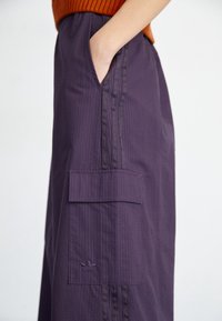 Lila cargo byxor med vertikala randiga mönster, sidofickor och en liten broderad logotyp. Tyget ser slätt ut med en lätt lyster.