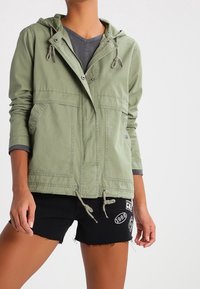 Grøn, let hættetrøjejakke med to forlommer, snøre i taljen og tryklåslukning. Stylet over en grå t-shirt og sorte shorts.