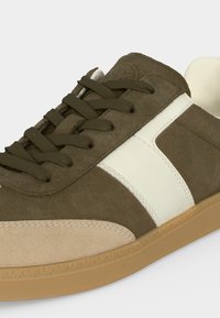 Χαμηλό sneaker με πράσινο σουέτ λαδί, μπεζ κάλυμμα στα δάχτυλα, λευκή πλαϊνή ρίγα, κορδόνια στο ίδιο χρώμα λαδί και καουτσούκ σόλα σε λευκό φόντο.