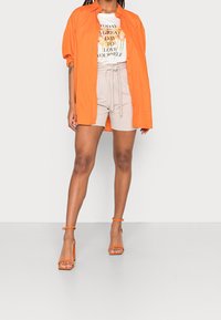 Henkilö, joka käyttää avointa oranssia paitaa, valkoista graafista t-paitaa, jossa on kukkateksti, beigeä vyöshortsia ja oransseja korokepohjaisia sandaaleja, seisoo valkoista taustaa vasten.