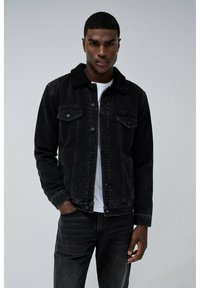 Chaqueta de mezclilla negra con un cuello forrado de forro polar, dos bolsillos en el pecho y cierres de botón, usada sobre una camisa blanca, combinada con unos vaqueros oscuros.