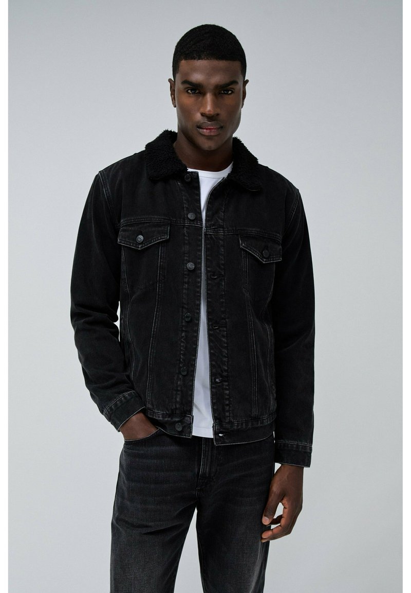 Chaqueta de mezclilla negra con un cuello forrado de forro polar, dos bolsillos en el pecho y cierres de botón, usada sobre una camisa blanca, combinada con unos vaqueros oscuros.