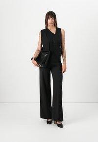 Svart skreddersydd vest med tre knapper, kombinert med vide bukser. Modellen holder en svart skinnveske og har på seg svarte høyhælte sko. Minimalistisk design.