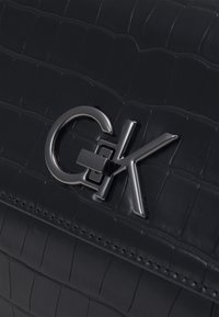 Czarna torba z eko-skóry o fakturze krokodyla, z metalową klamrą z logo w srebrnym kolorze. Szwy widoczne wzdłuż krawędzi.