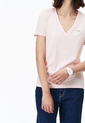 Kobieta w jasnoróżowej koszulce z dekoltem w serek, ciemnoniebieskich dżinsach oraz białym zegarku na nadgarstku lewej ręki, na białym tle.