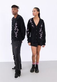 Μαύρα πουλόβερ με ροζ σχέδιο που θυμίζει φλόγες, με κουμπιά και μακριά μανίκια, φορεμένα με φθαρμένα μαύρα παντελόνια και μποτάκια στρατιωτικού στυλ.