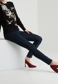 Černý svetr s květinovou výšivkou, modré skinny džíny a červené vysoké podpatky. Oblečení se vyznačuje hladkými texturami a obtaženými siluetami.