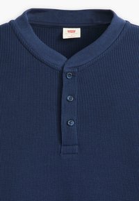 Tengerészkék henley póló bordázott anyagból, három kék gombbal ellátott gombos nyakkal és kerek gallérral. 