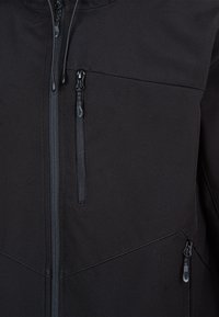 Giacca softshell nera con diverse tasche zip, tessuto testurizzato e design elegante con cuciture curve e cordini regolabili.