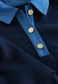 Granatowy dzianinowy polo z jasnoniebieskim denimowym kołnierzykiem. Posiada trzy okrągłe złote guziki. Miękka tekstura, z prążkowanymi mankietami i czystym designem.