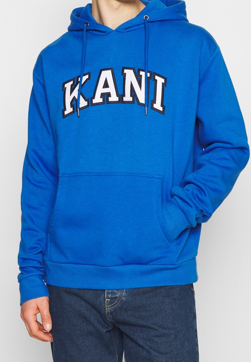 Sudadera con capucha de algodón azul con la palabra "KANI" bordada en letras blancas y negras. Cuenta con un bolsillo delantero y capucha con cordón ajustable.