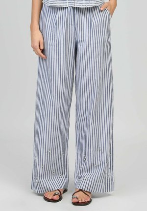 Femme portant un pantalon large à rayures verticales bleu et blanc avec de petites perles décoratives près de l'ourlet, accompagné de sandales noires.