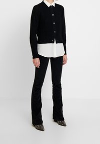 Schwarze gerippte Strickjacke mit Knöpfen, getragen über einem weißen Hemd, kombiniert mit schwarzen ausgestellten Hosen und Schlangenleder-Print-Stiefeletten mit Absätzen.