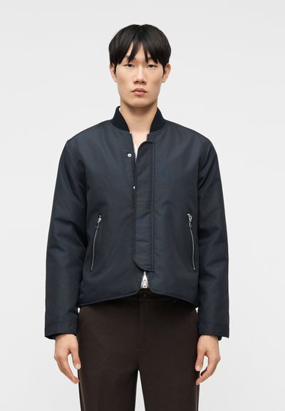 Jeune homme portant une veste bomber noire zippée avec des poches à fermeture éclair et un pantalon marron foncé sur un fond blanc uni.