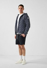 Uomo con giacca con cappuccio blu navy a motivo, t-shirt bianca, pantaloncini neri, calzini bianchi e scarpe da ginnastica bianche, in piedi contro uno sfondo bianco uniforme.