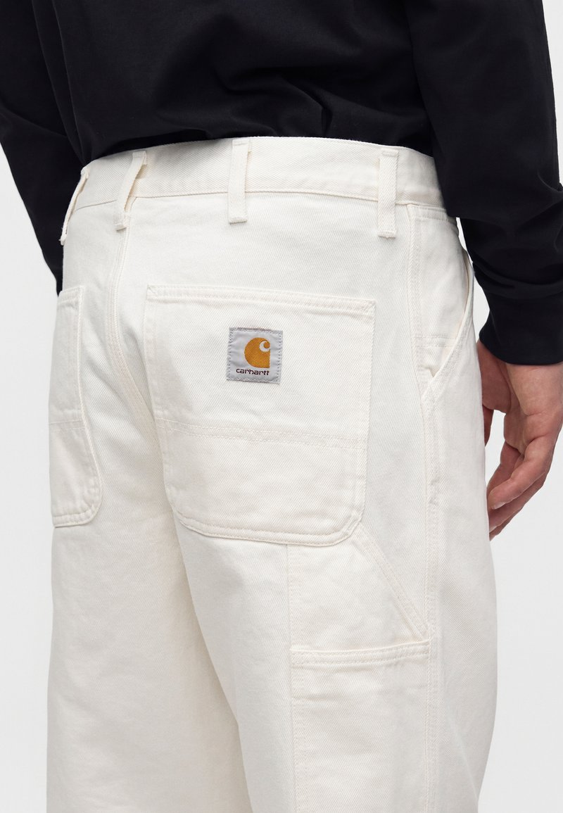 Pantalon en denim blanc avec grandes poches arrière et latérales, porté par une personne en chemise noire à manches longues, montrant le logo Carhartt sur la poche arrière.