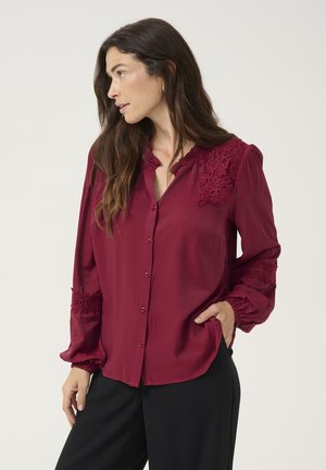 Burgundy blouse with a button front, lace accents on shoulders and sleeves, and elastic cuffs, featuring a smooth, lightweight fabric.