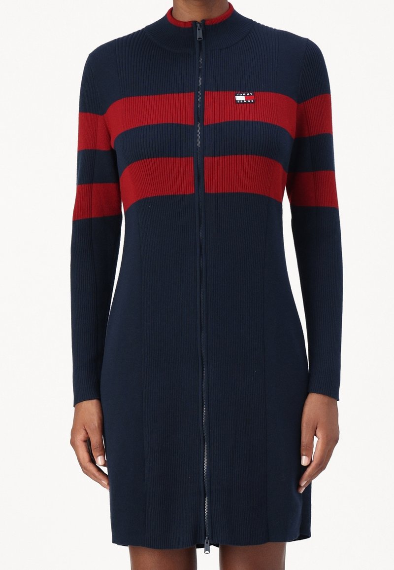 Robe en maille côtelée marine avec des rayures horizontales rouges sur la poitrine et les manches, fermeture éclair intégrale à l'avant et petite étiquette logo sur la poitrine gauche.