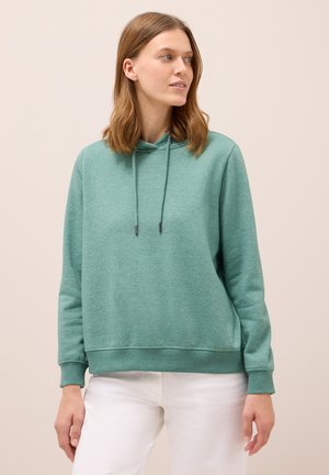 Frau trägt einen grünen Kapuzenpulli mit Kordelzügen und weiße Hose, schaut leicht nach rechts vor einem schlichten Hintergrund.