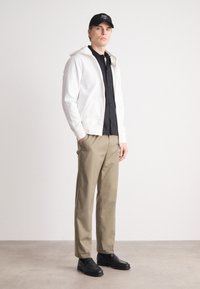 Felpa bianca con zip sopra una camicia nera a bottoni, abbinata a un paio di pantaloni khaki e scarpe nere. Design semplice, vestibilità comoda, minimi dettagli.