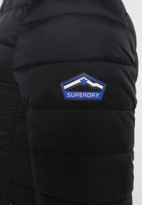 Sort dunjakke med quiltede sektioner, et fremtrædende blåt og hvidt "Superdry" logo-patch på venstre bryst og en glat, skinnende overflade.