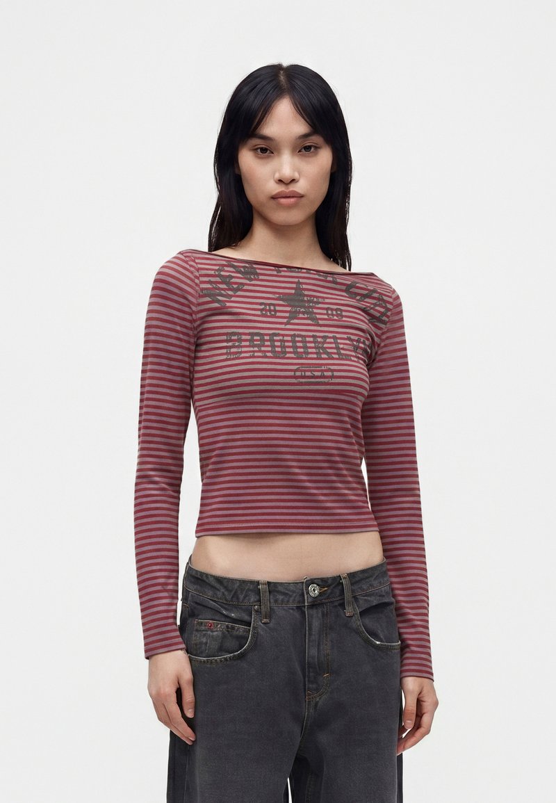 Γυναίκα φοράει κόκκινο και γκρι ριγέ crop top με μακριά μανίκια και κείμενο, καθώς και ξεθωριασμένο μαύρο τζιν, μπροστά σε απλό φόντο.