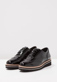 Brogues en cuir verni noir avec des détails perforés, bout rond, devant à lacets et semelle empilée avec des couches marron et noires.