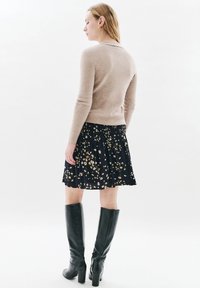 Nainen, jolla on vaaleanbeige villapaita, tummansininen kukkamekko ja mustat polven yli ulottuvat saappaat, seisoo sivuttain valkoista taustaa vasten.