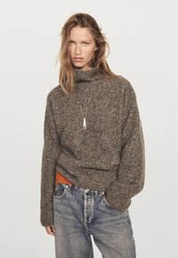 Donna che indossa un maglione spesso grigio, jeans blu e una collana con pendente, su uno sfondo leggero e semplice.