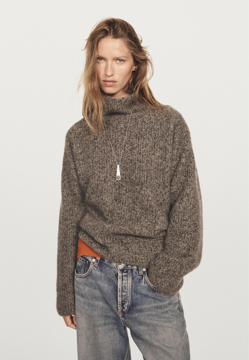 Donna che indossa un maglione spesso grigio, jeans blu e una collana con pendente, su uno sfondo leggero e semplice.