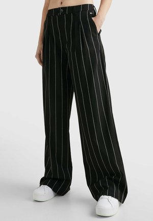 Czarne spodnie o szerokich nogawkach z białymi pionowymi prążkami, noszone z białymi platformowymi tenisówkami i rękami w kieszeniach.