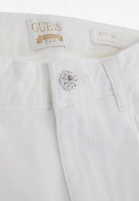 Pantalon en denim blanc avec fermeture à bouton ornée de cristaux, taille haute et logo GUESS sur la ceinture.
