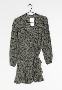 Grünes Kleid mit schwarzem Punktenmuster, Rüsche an der Vorderseite und an den Ärmelbündchen, mittellang, leichter Stoff, hängt an einem schwarzen Haken.