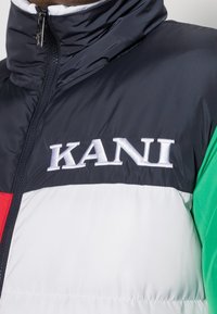 Kurtka puchowa z wysokim kołnierzem, w kolorze blokowym w odcieniach granatu, bieli, czerwieni i zieleni, z haftem "KANI" na klatce piersiowej.