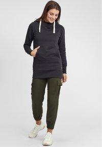Felpa con cappuccio grigio scuro dotata di tasca frontale e cordini beige, abbinata a pantaloni cargo oliva e scarpe da ginnastica bianche.
