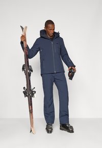 Marineblå ski-jakke med hette og glidelåser, parret med matchende bukser. Personen holder ski og skibriller, iført svarte skistøvler.