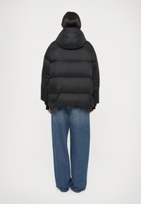 Geacă puffer neagră cu glugă, având un design pătrățos și textură matlasată. Asortată cu blugi de denim albastri, cu croială largă.