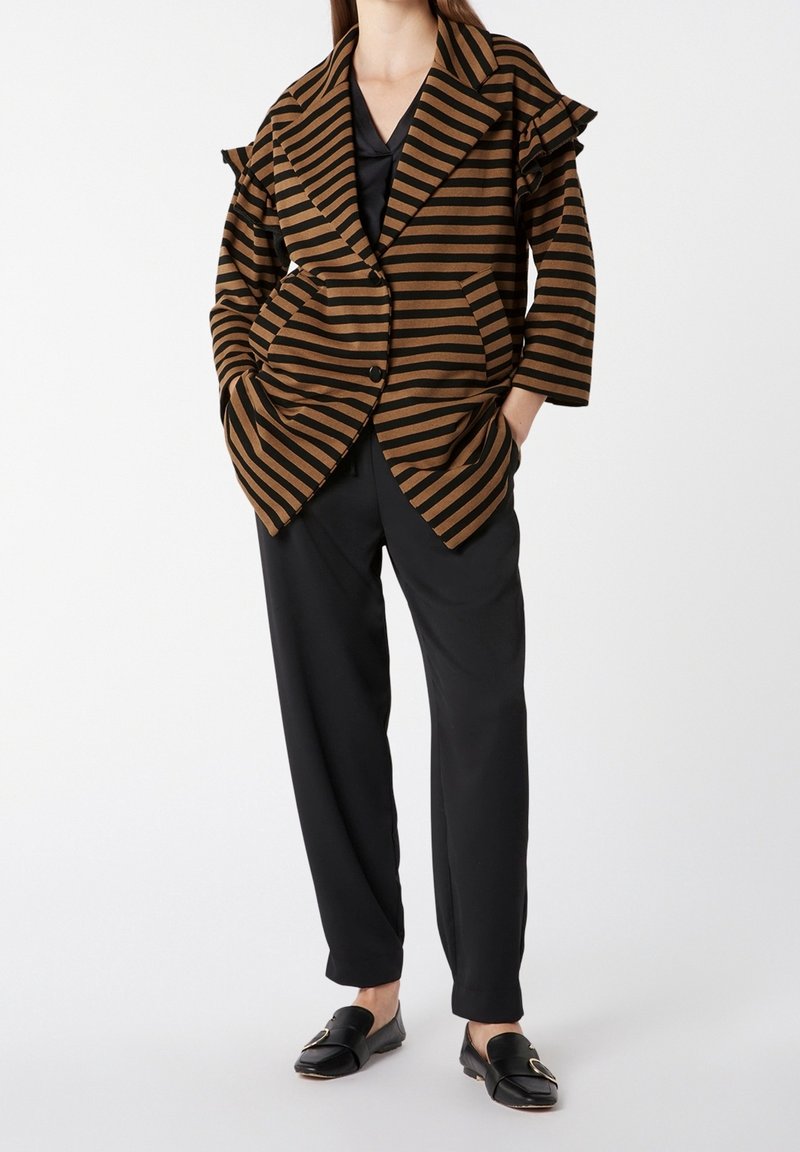 Femme portant un blazer oversize rayé marron et noir avec des détails à volants sur les épaules, un pantalon noir et des mocassins noirs sans lacets.