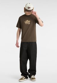 Bruin katoenen T-shirt met gele "VANS" logo, gecombineerd met losse zwarte denim broek en bruin-witte sneakers. Grijze en beige pet.