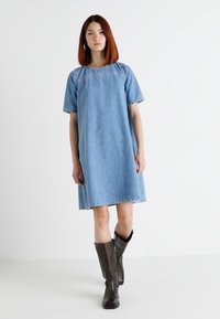 Gaiši zils džeza kleita ar īsām piedurknēm, apaļu kakla izgriezumu un taisnu siluetu. Saskaņota ar tumši brūnām ceļgala garuma zābakiem.