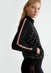 Maglione nero in maglia con motivo testurizzato del logo, colletto alto e zip. Presenta accenti a righe rosse e metalliche in contrasto sulle maniche.