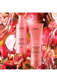 Två Aveda Nutri Plenish-flaskor: en rund rosa schampo-flaska och en rosa tubbalsam, omgivna av röda frukter och växtelement.