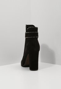 Botín de ante negro con tacón grueso, que presenta detalles de tachuelas y una banda ancha alrededor del tobillo. Textura suave, diseño minimalista.