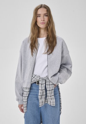 Fiatal, hosszú hajú nő szürke cipzáras dzsekiben, fehér ingben, deréknál kötött kockás inggel és kék farmerral, egyszerű háttér előtt.