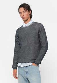 Grau gestrickter Pullover mit einem strukturierten Muster und geripptem Kragen, getragen über einem blau-weiß gestreiften Hemd, kombiniert mit hellblauen Jeans.