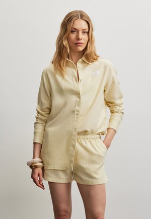 Kvinde med bølget blondt hår iført en lys gul knap-op skjorte og matchende shorts, stående med den ene hånd i lommen og armbånd på håndleddet.