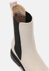 Bottine en cuir beige avec côtés élastiques noirs, un intérieur texturé tan, et une semelle en caoutchouc noir robuste. Équipée d'une languette à l'arrière.